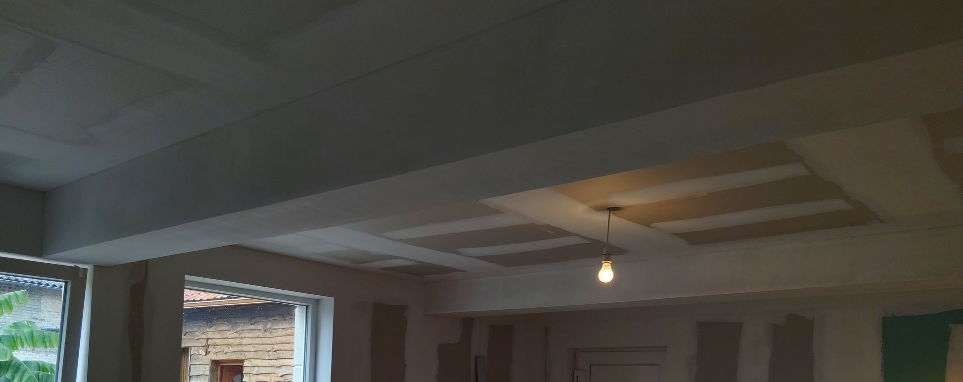 Pièce intérieure avec des poutres apparentes au plafond, une ampoule unique et des fenêtres ouvertes.