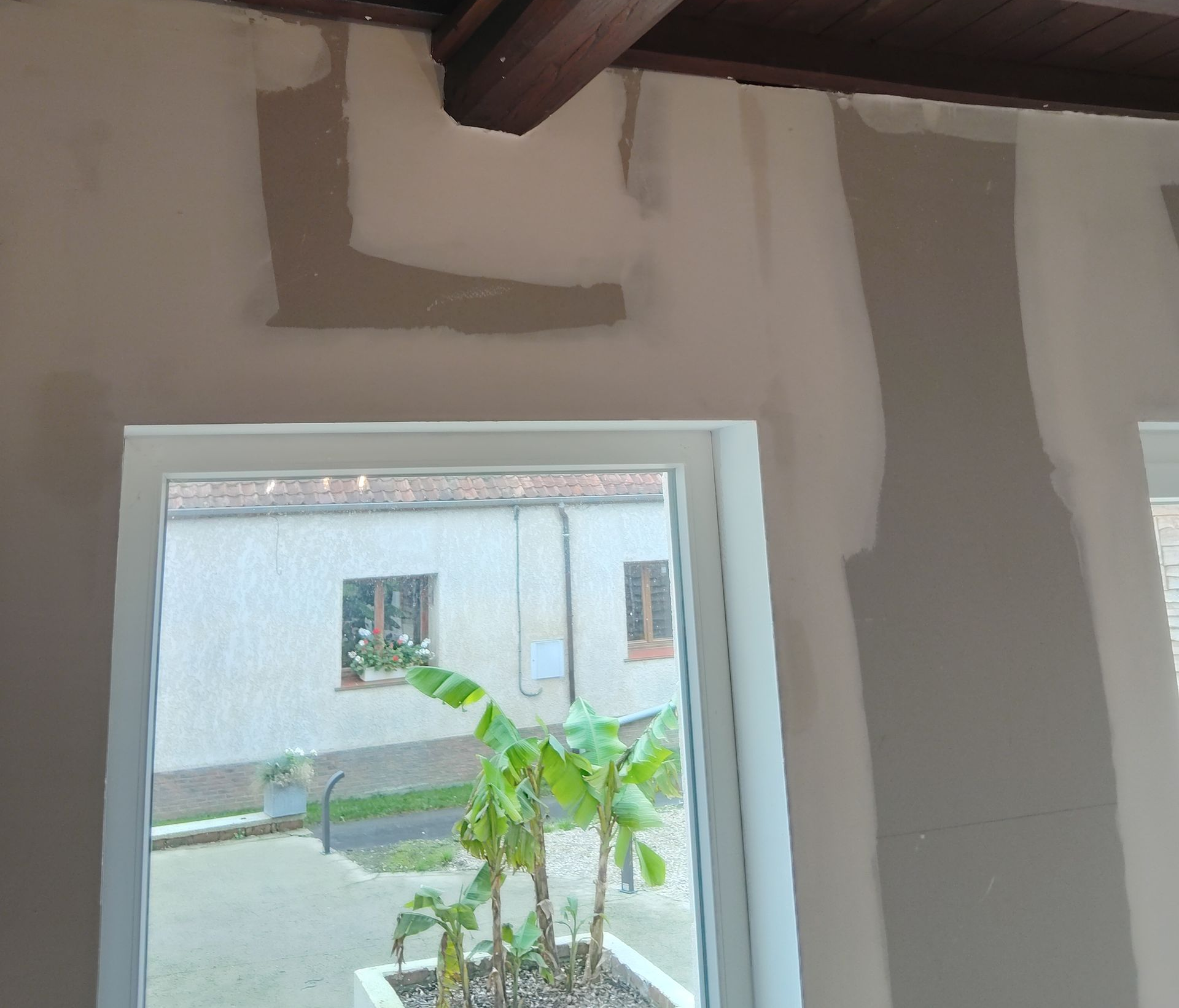 Mur intérieur en cours de rénovation autour d’une fenêtre, avec vue sur un petit bananier à l’extérieur.