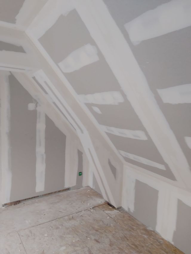 Pièce de grenier en cours d’aménagement, avec des plaques de plâtre non terminées, des joints recouverts de bandes, un plafond en pente et un sol nu.