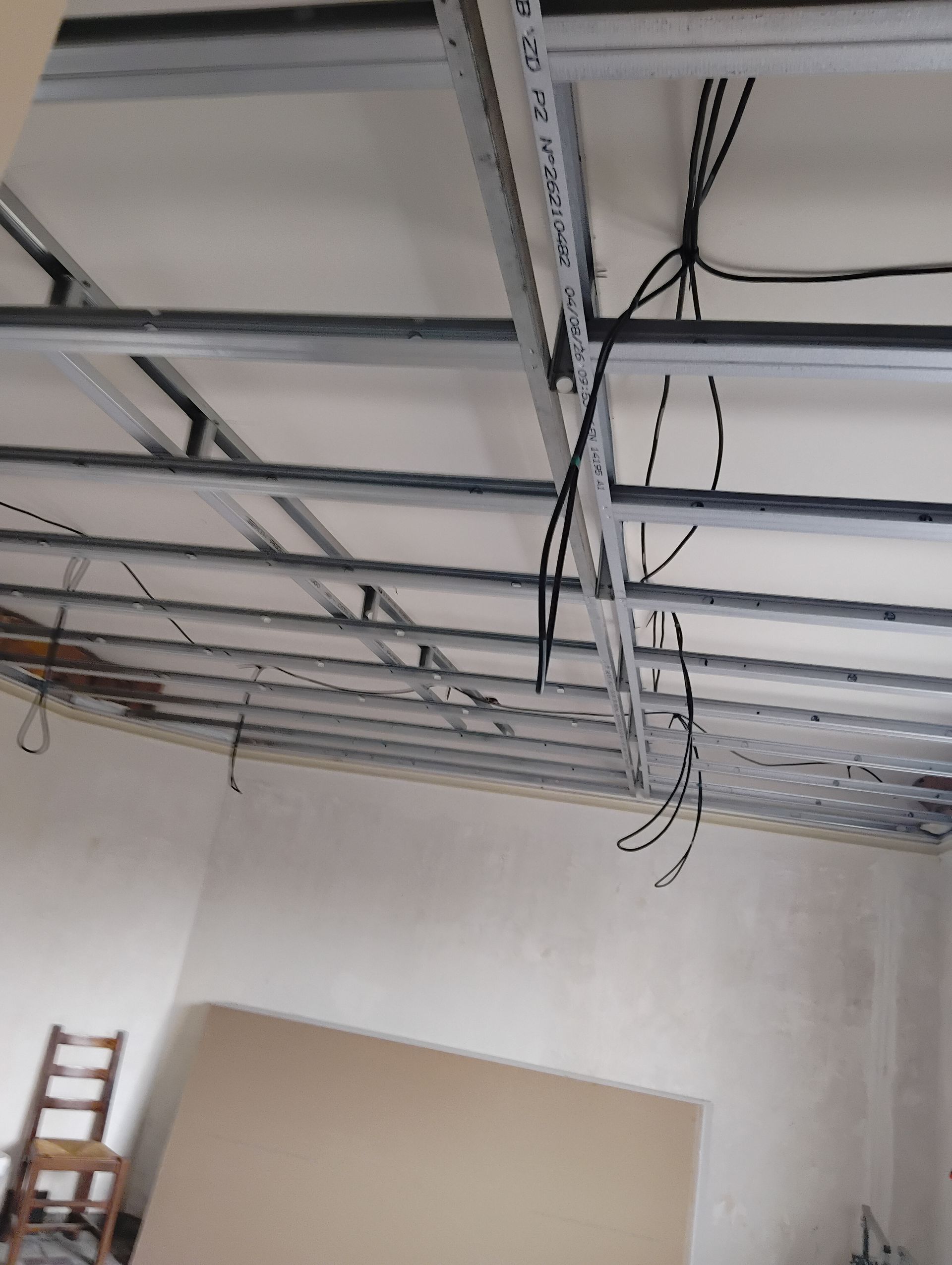 Plafond de garage avec des rails apparents et des fils suspendus, au-dessus d’un mur partiellement peint et d’une échelle.