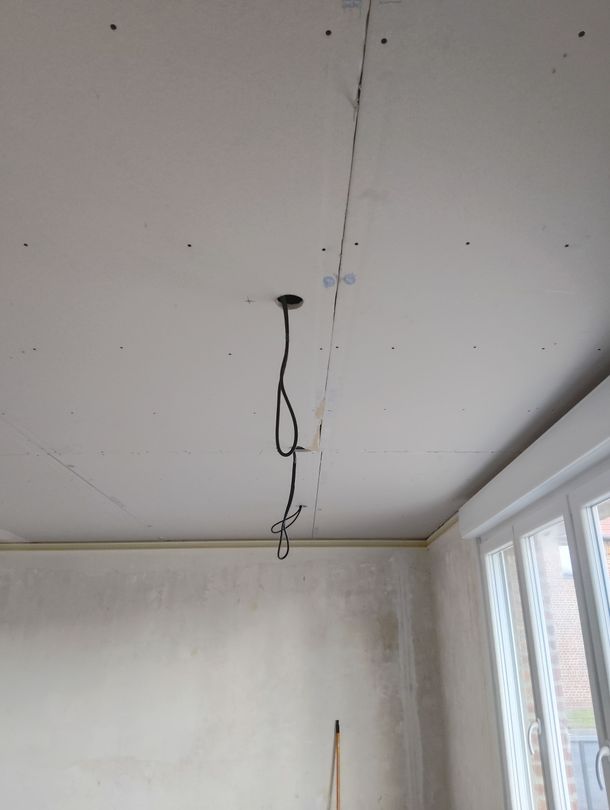 Pièce vide avec un plafond, des fils électriques pendants et une fenêtre laissant entrer la lumière sur la droite.