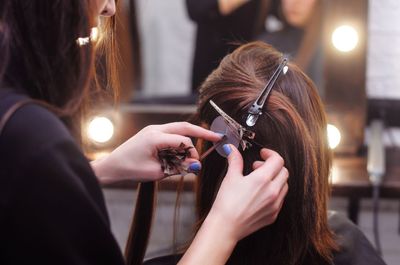Eine Friseurin benutzt eine kleine Plastikscheibe und eine Metallklemme, um braunes Haar in einem Salon mit Schminkspiegelbeleuchtung abzuteilen.