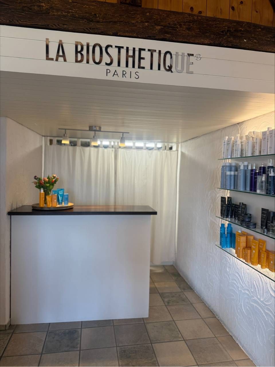 Ein sauberer Empfangsbereich für einen La Biosthetique Paris Salon mit weissem Tresen, Produkten in Regalen und Fliesenboden.