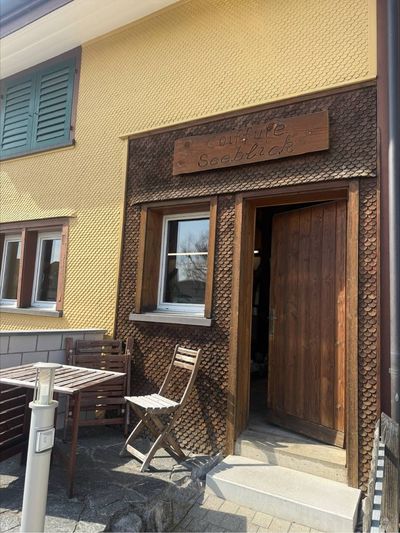 Ein Eingangsbereich mit Holztür und kleinem Fenster neben einem Tisch und einem Stuhl auf einer Steinterrasse vor einem gelben Gebäude.