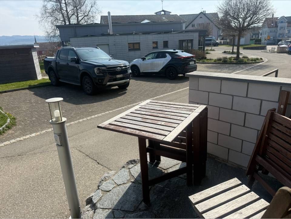 Eine Terrasse im Freien mit einem hölzernen Klapptisch und Stuhl in der Nähe eines Parkplatzes mit zwei Autos unter klarem, sonnigem Himmel.