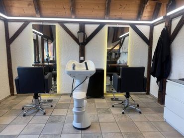 Ein Friseursalon mit zwei schwarzen Frisierstühlen, einem zentralen weissen Waschbecken, grossen vertikalen Spiegeln und Holzbalken.