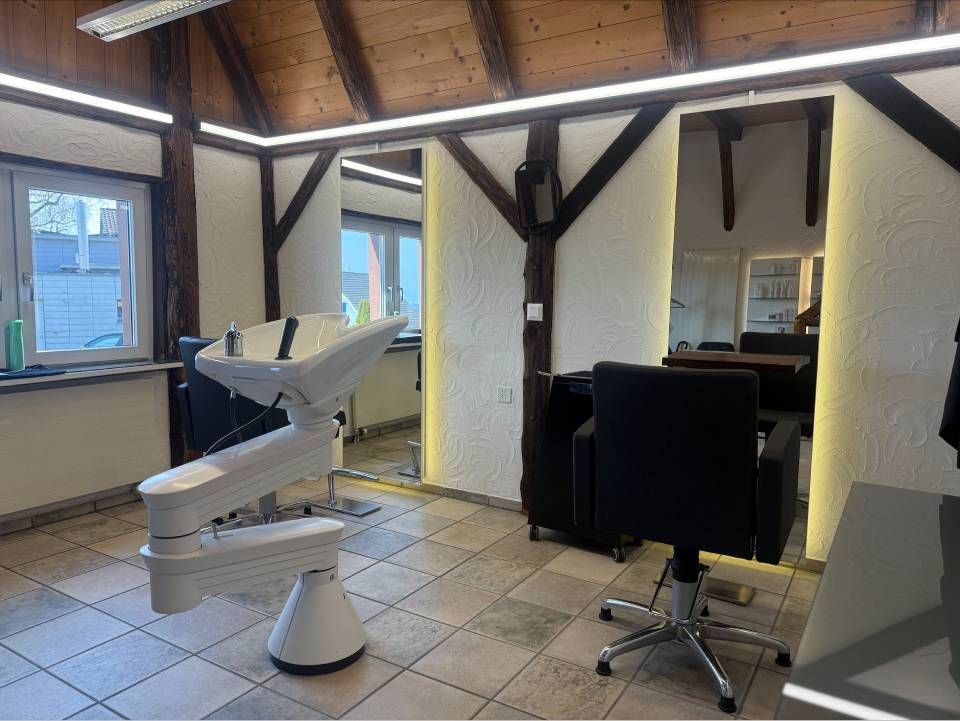 Ein heller Friseursalon mit weissem Waschbecken und schwarzem Friseurstuhl vor einem Spiegel.