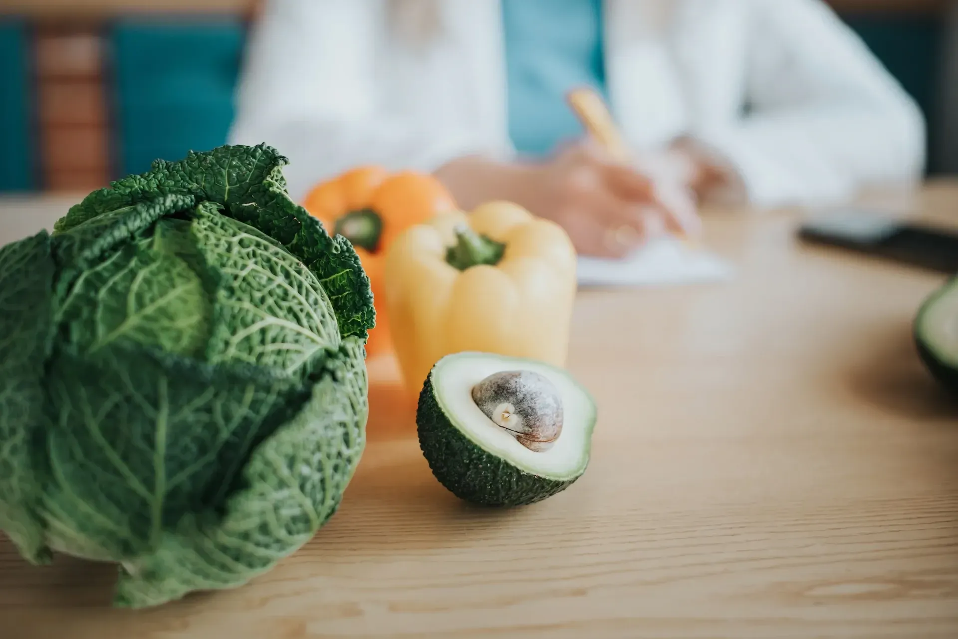 Primer plano de verduras frescas, como repollo, pimientos y aguacate, sobre una mesa con una persona escribiendo al fondo.