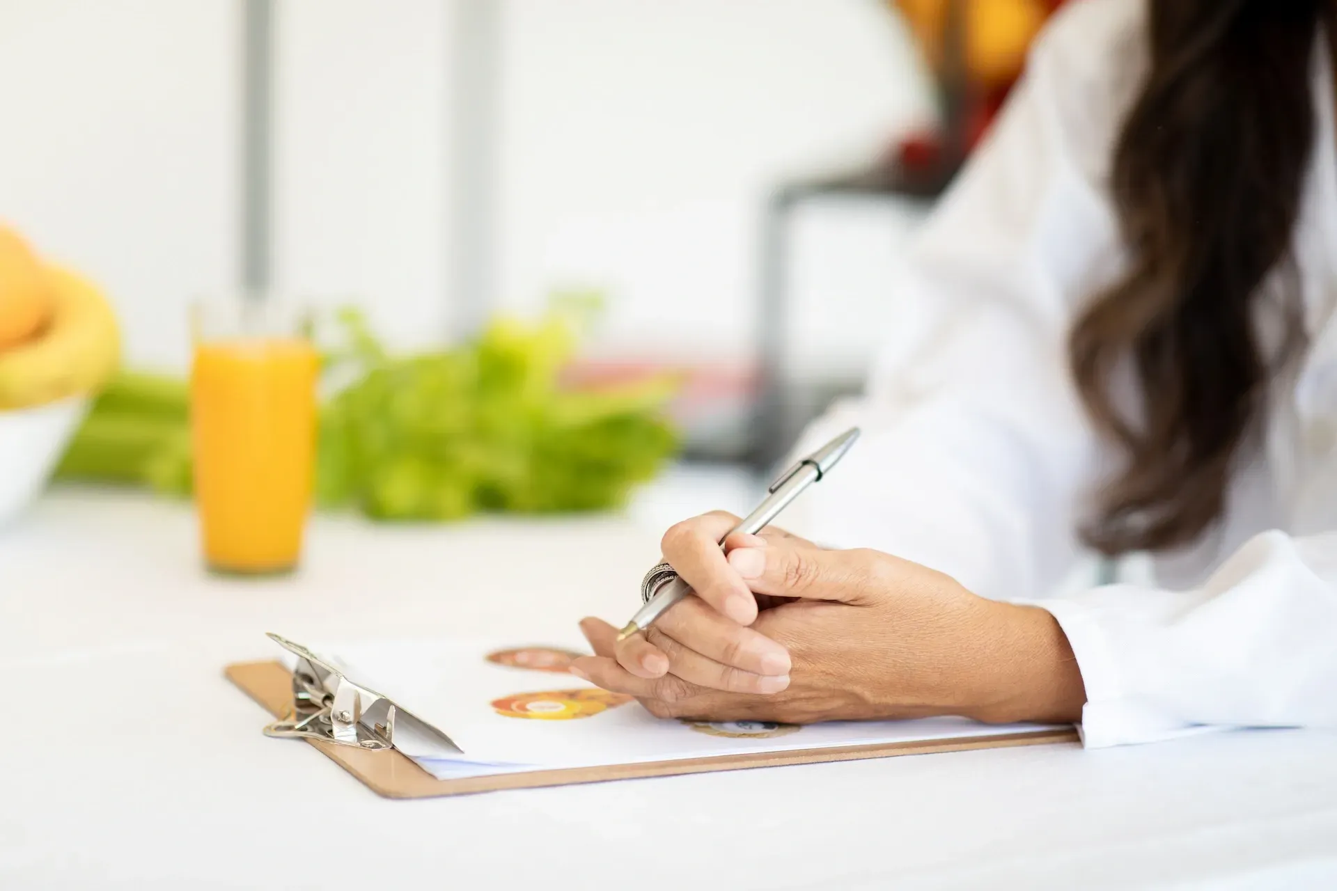 Persona con bata blanca escribiendo en un portapapeles, con fruta y jugo cerca.