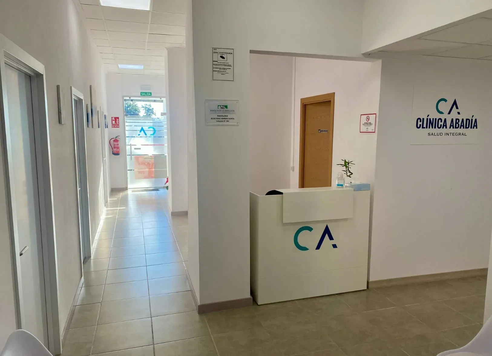 Pasillo largo en una clínica con recepción, puertas y una luz brillante al final. Se ve el logo 
