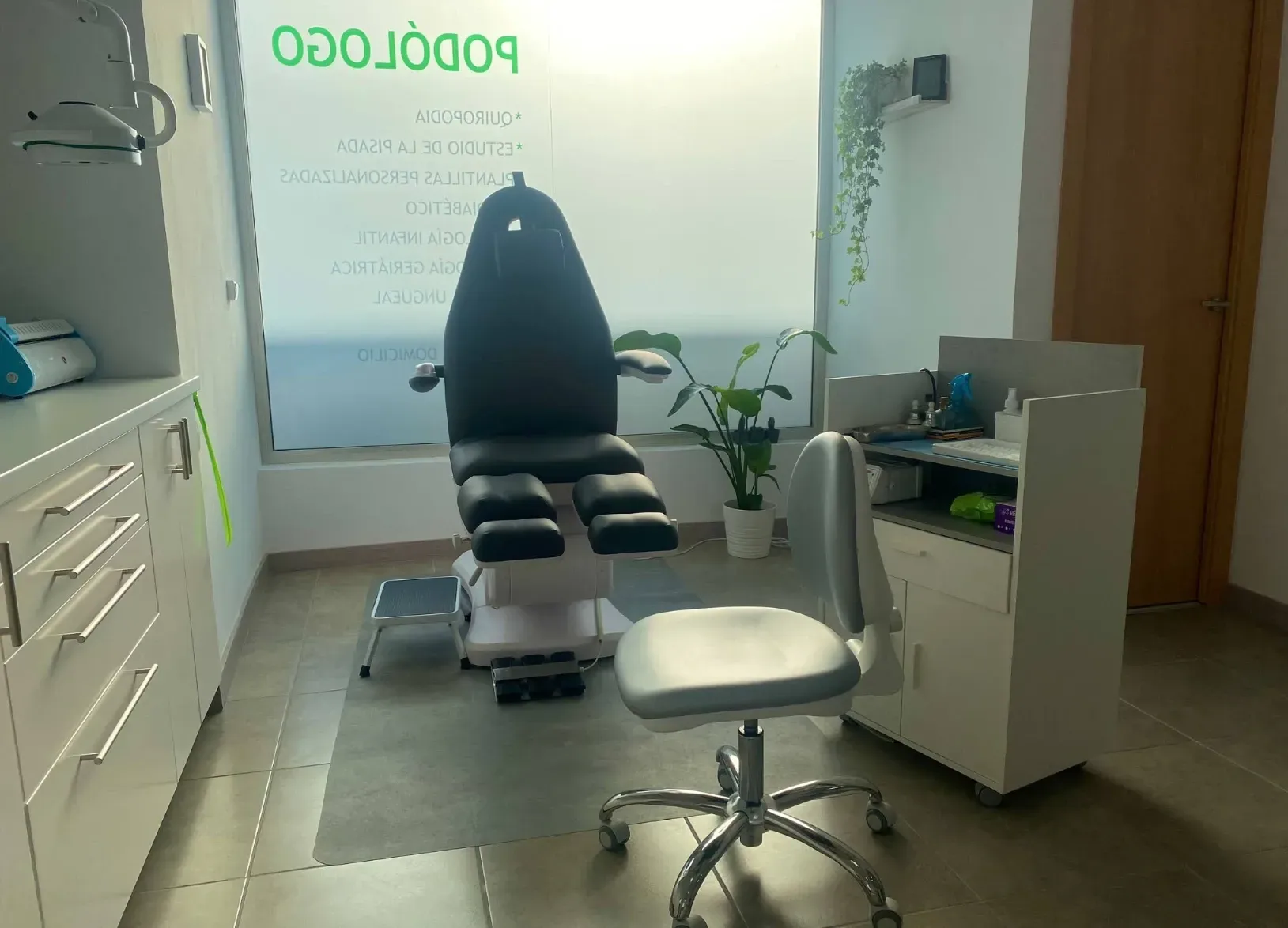 Interior de clínica de podología: sillón de exploración, armarios blancos, pequeño taburete.