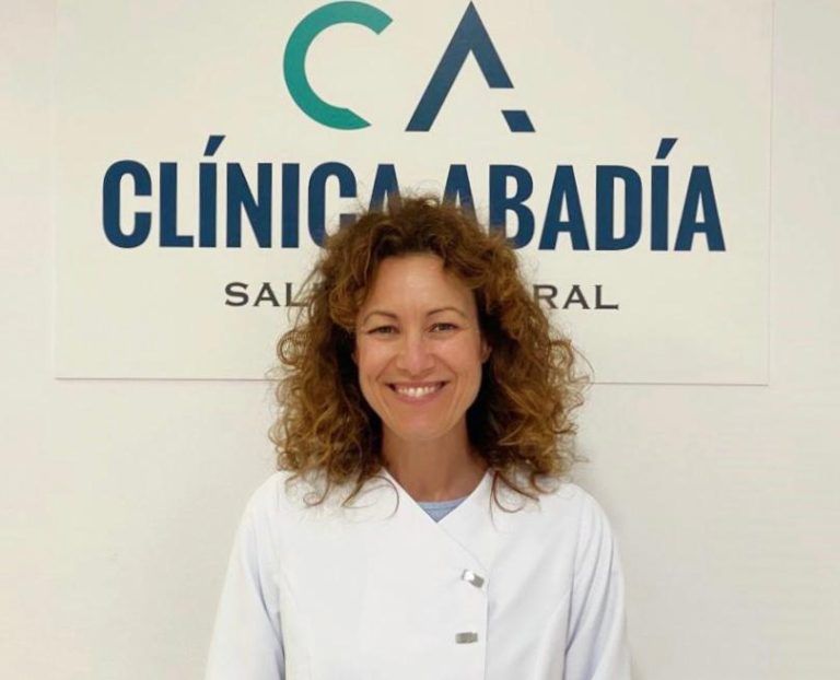 Mujer sonriente con bata blanca, frente al cartel de 