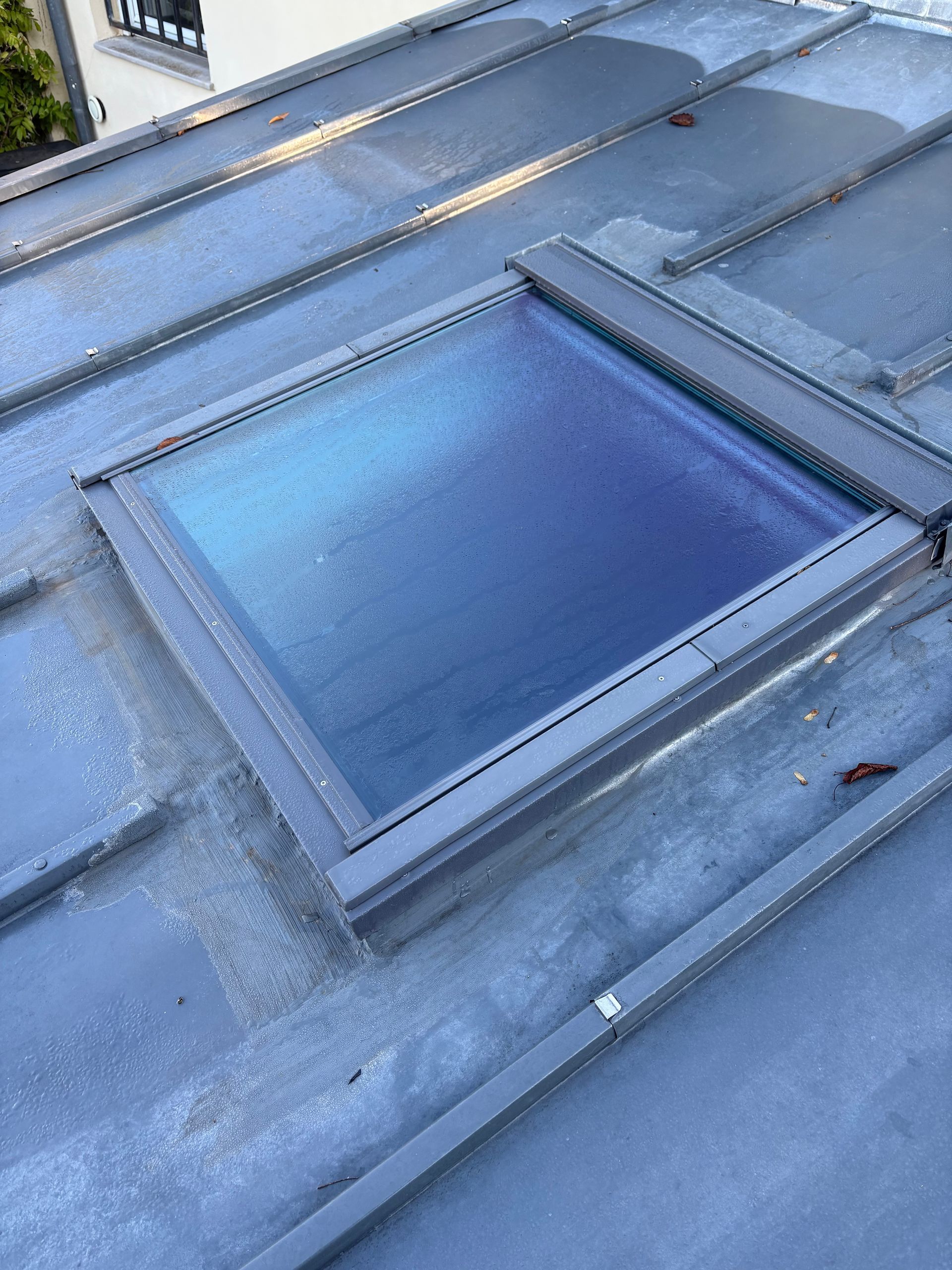 Puits de lumière sur un toit en métal gris, possiblement humide, avec un cadre de fenêtre.