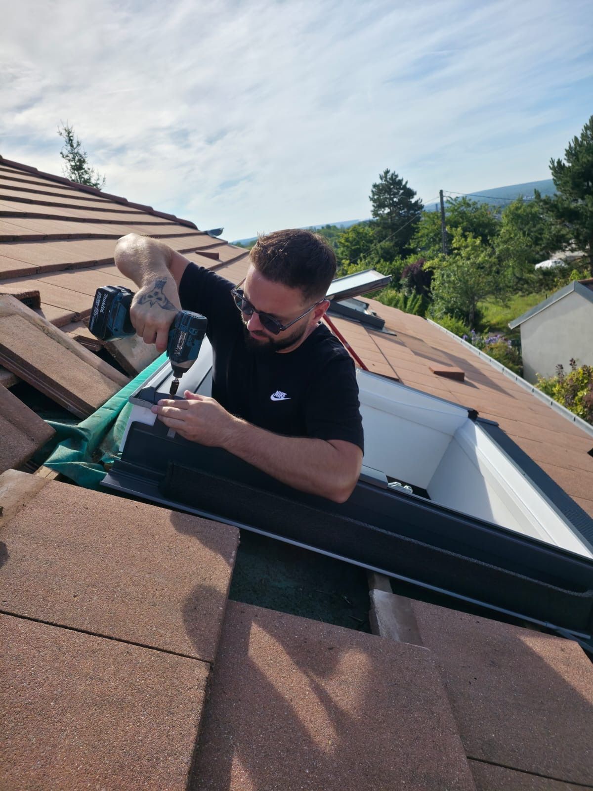 Un homme portant des lunettes de soleil visse un cadre de puits de lumière sur un toit de tuiles. Journée ensoleillée.
