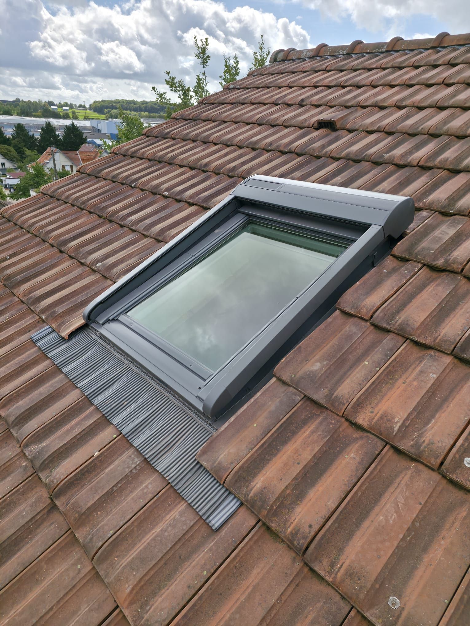 Puits de lumière installé sur un toit de tuiles brunes, sur fond de ciel nuageux.