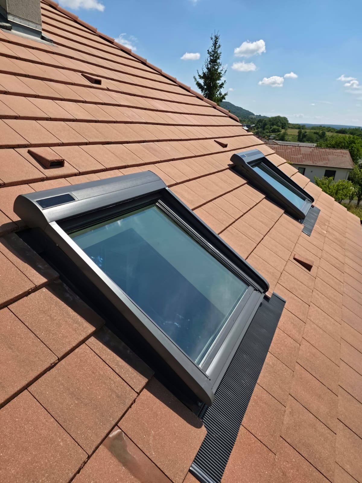 Deux puits de lumière sur un toit de tuiles sous un ciel bleu, avec vue sur des arbres verts en arrière-plan.