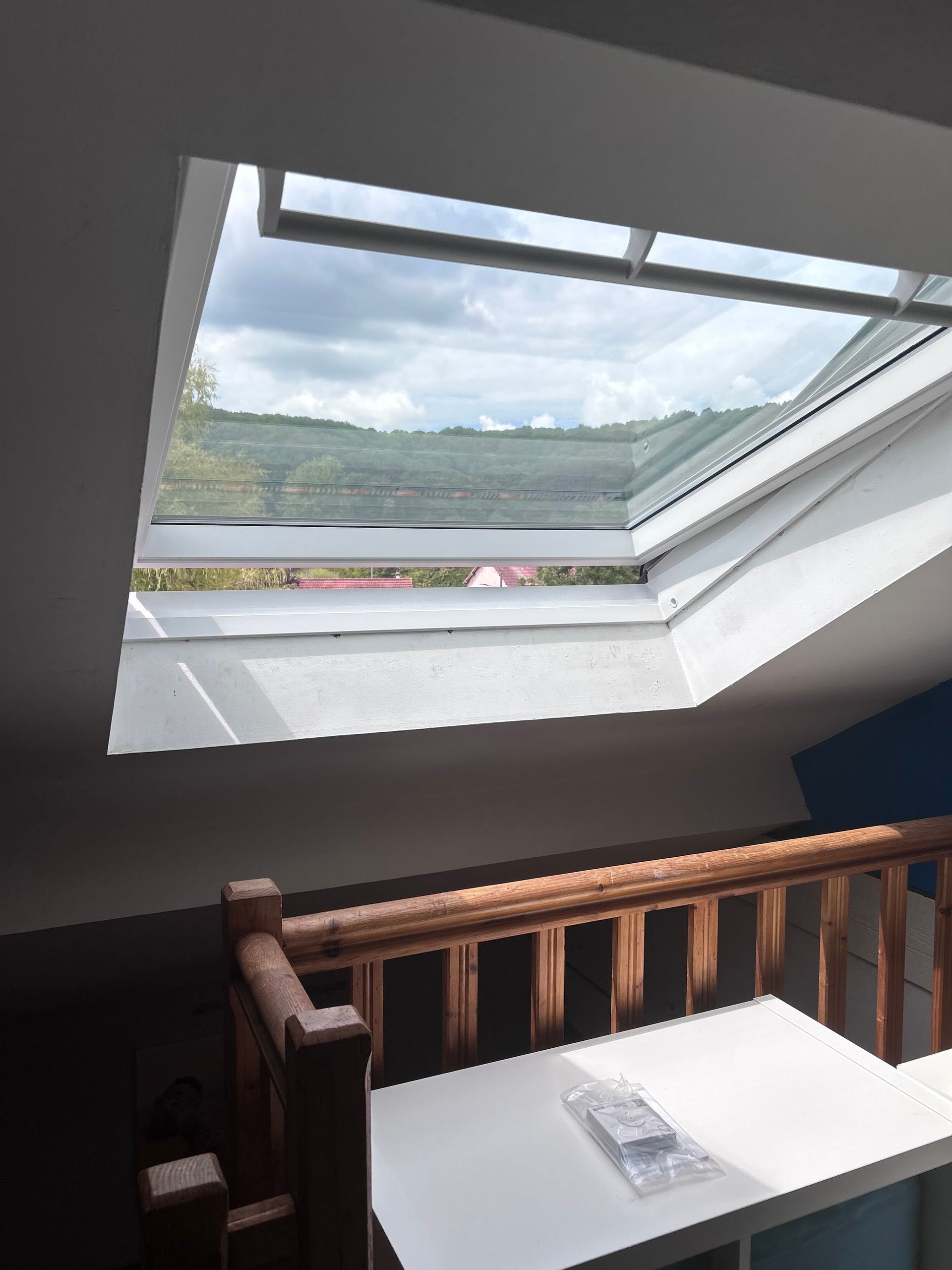 Fenêtre de toit dans une pièce donnant sur des collines verdoyantes. Table blanche et balustrade en bois au premier plan.