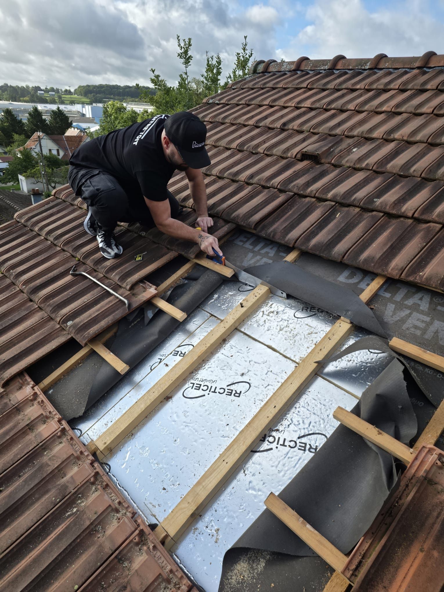 Un couvreur, agenouillé sur un toit de tuiles, découpe des matériaux. On aperçoit les poutres et l'isolant. Ciel nuageux.