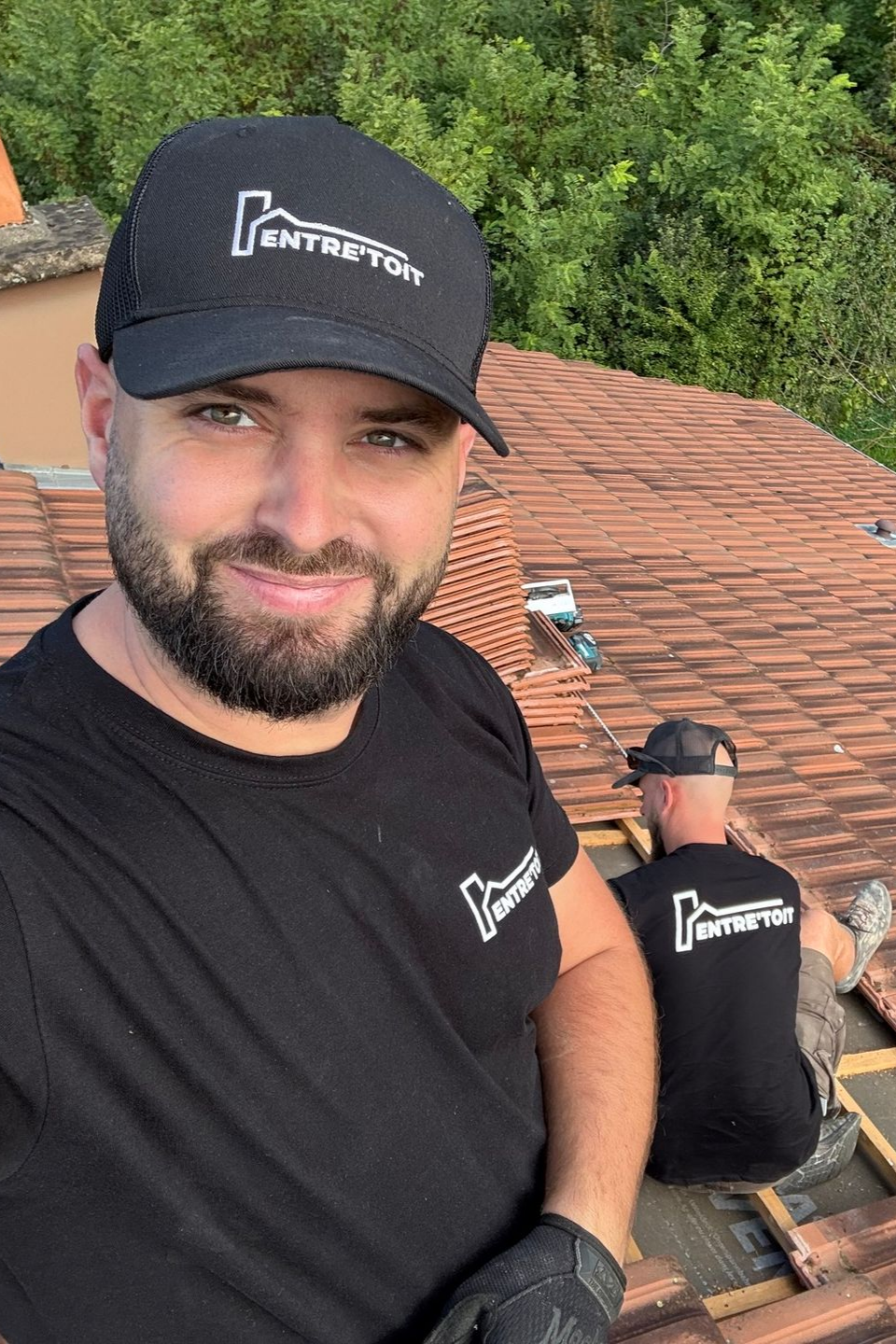 Deux hommes sur un toit de tuiles, l'un prenant un selfie, portant des casquettes et des chemises Entretoit noires.