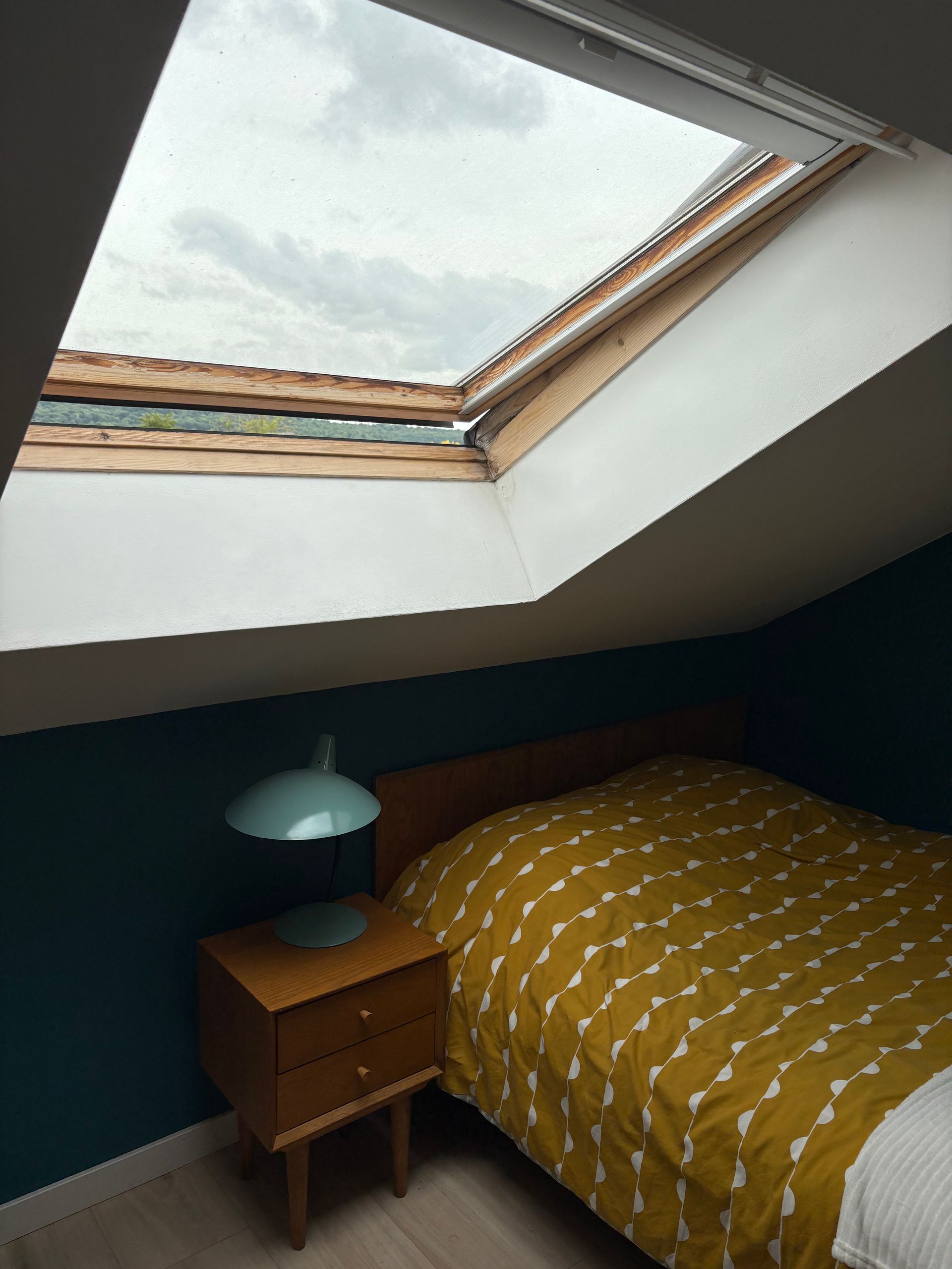 Chambre avec puits de lumière, lit recouvert d'une couverture jaune, lampe et mur turquoise. Ciel nuageux visible.