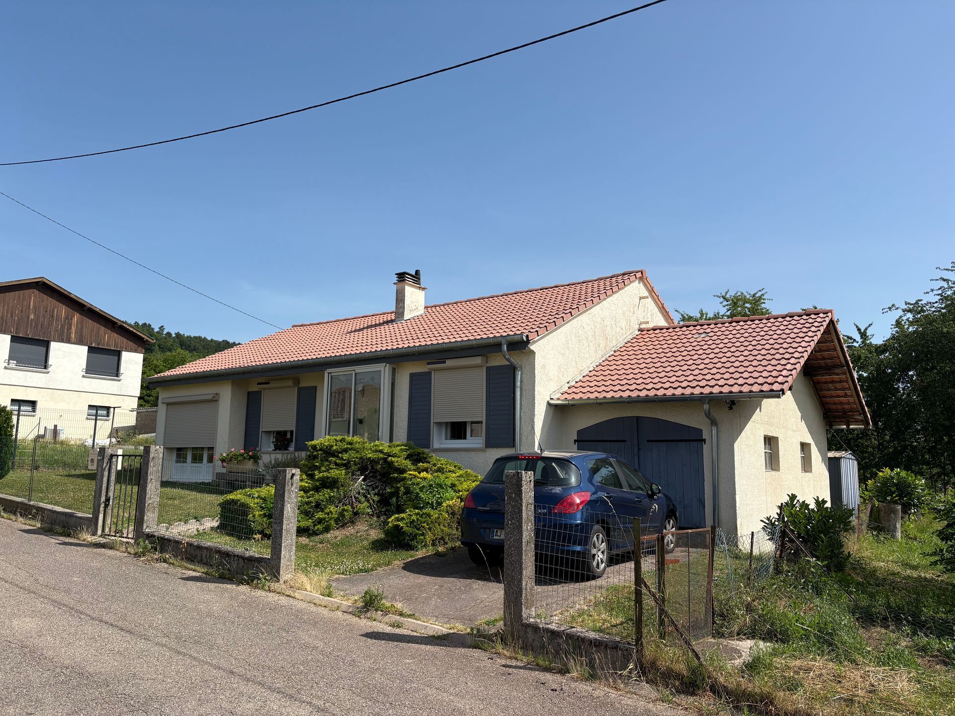 Une petite maison claire au toit de tuiles. Une voiture bleue est garée dans l'allée.
