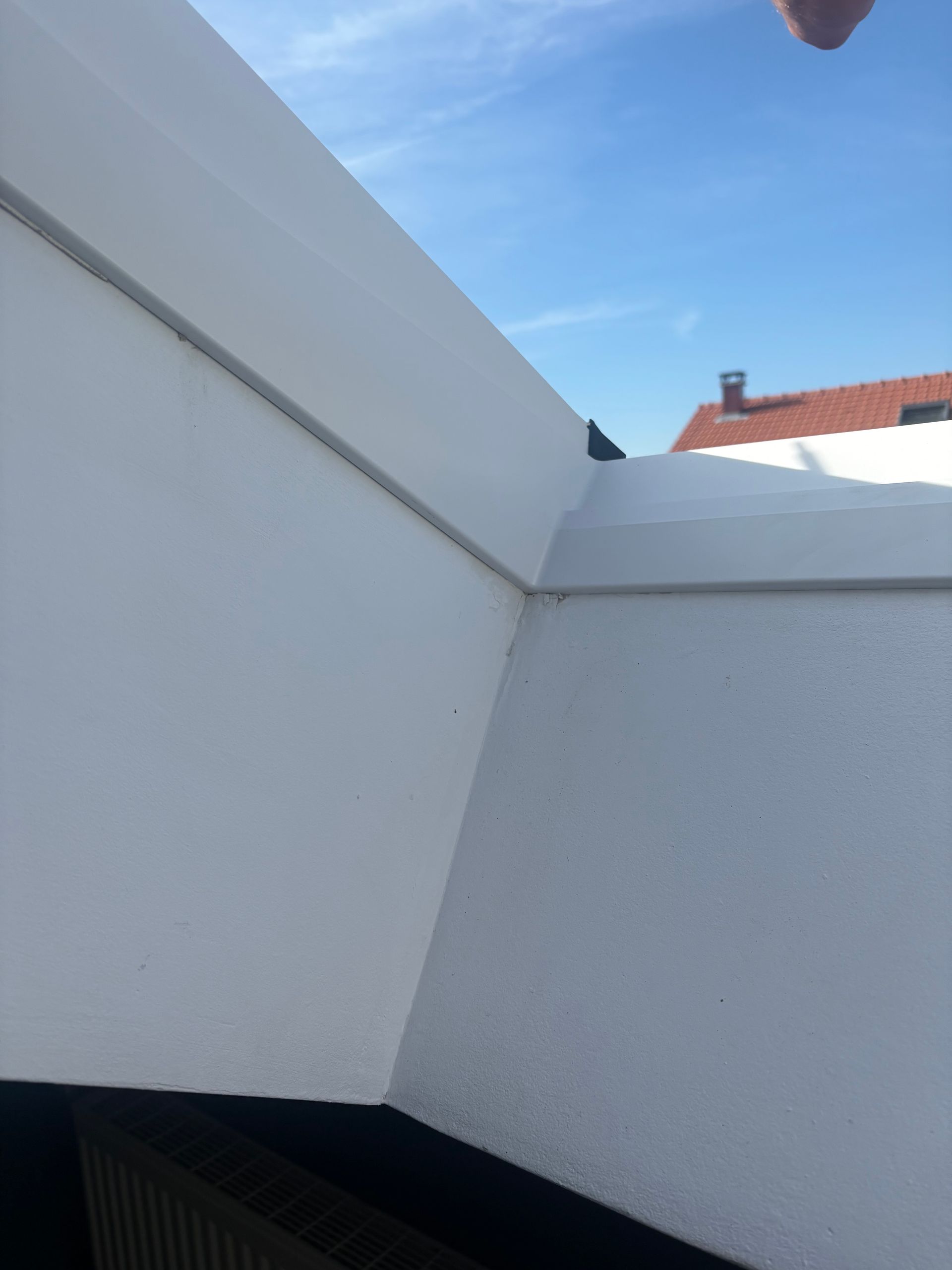 Un coin de toit peint en blanc se détachant sur un ciel bleu, avec un bâtiment au toit rouge en arrière-plan.