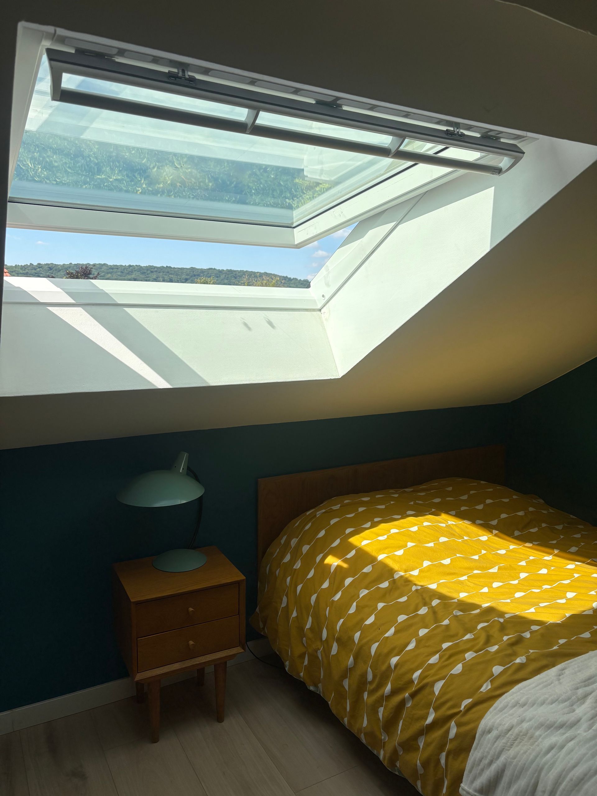Puits de lumière dans une chambre ouverte sur la verdure. Lit avec couette à motifs jaunes, table de chevet et mur turquoise.