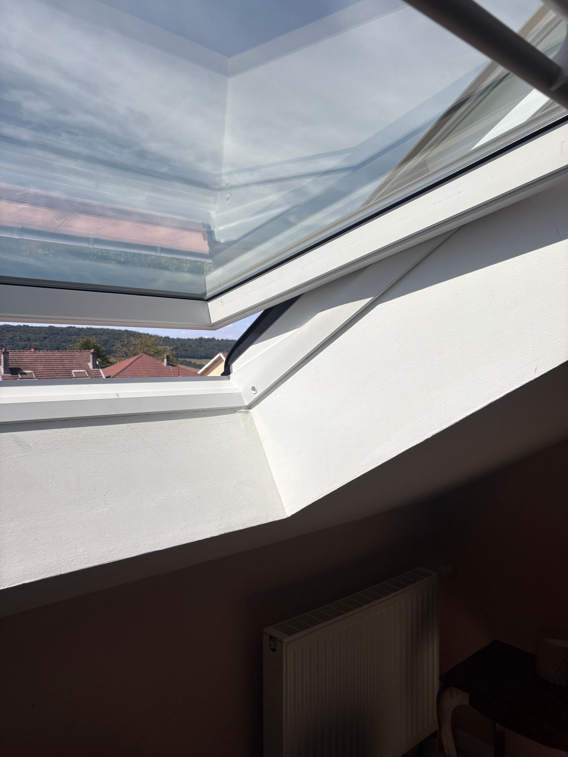 Une lucarne à cadre blanc est ouverte, offrant une vue sur les toits et le ciel.