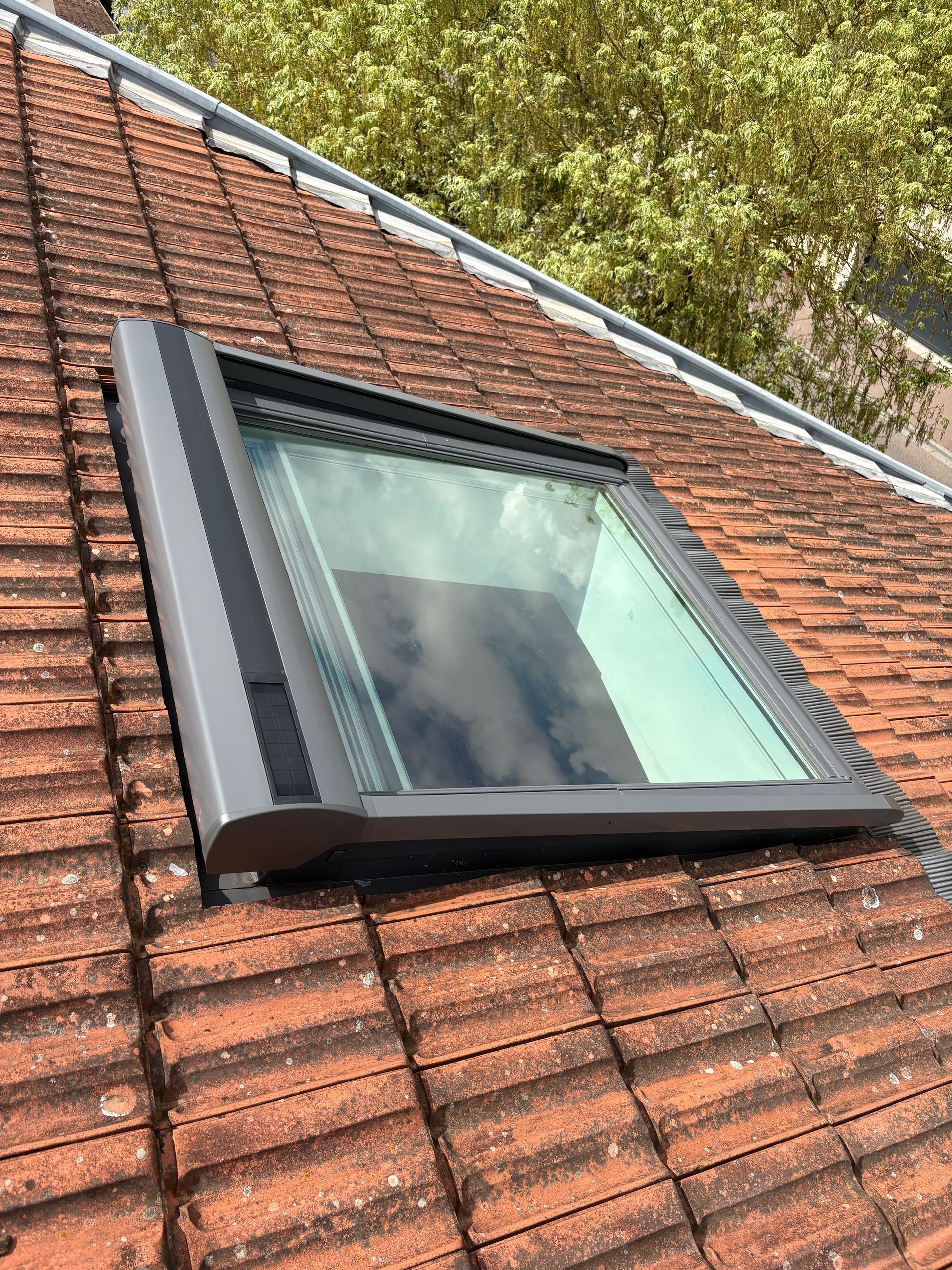 Puits de lumière sur un toit de tuiles rouges, encadré de métal sombre. 