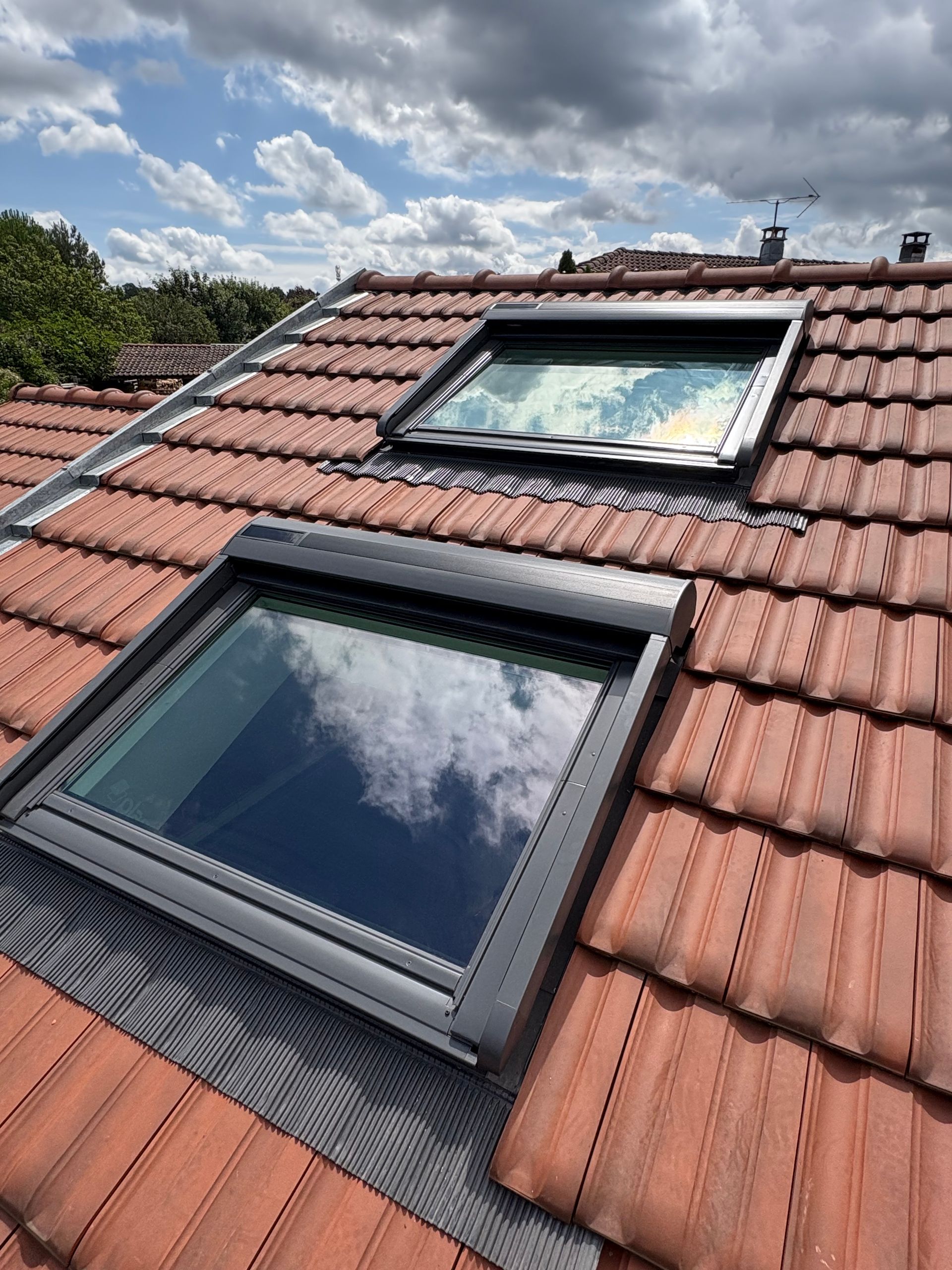 Deux puits de lumière sur un toit de tuiles rouges reflètent un ciel nuageux.