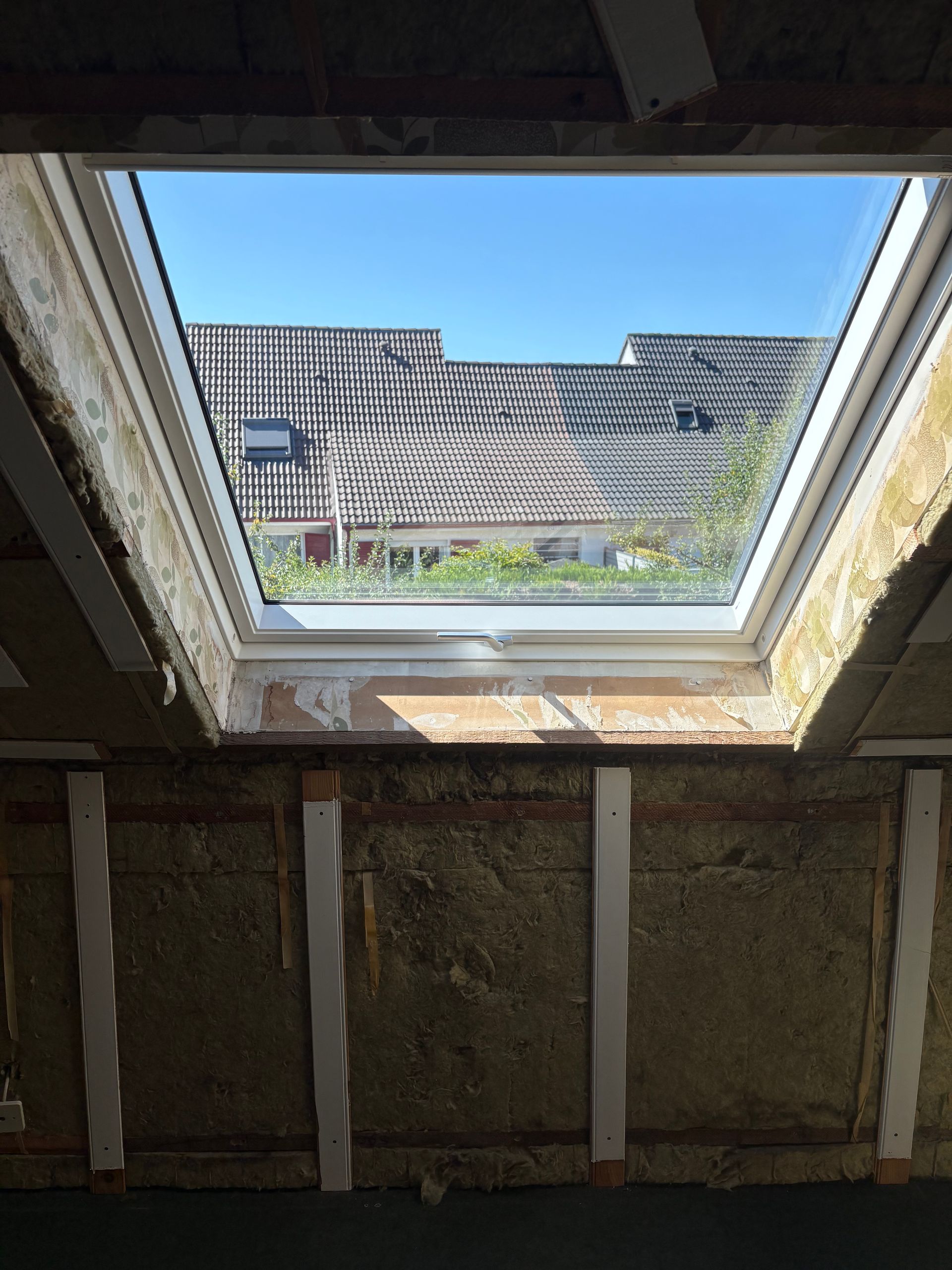 Puits de lumière dans une pièce, offrant une vue sur les toits et le ciel. Construction intérieure avec isolation apparente.