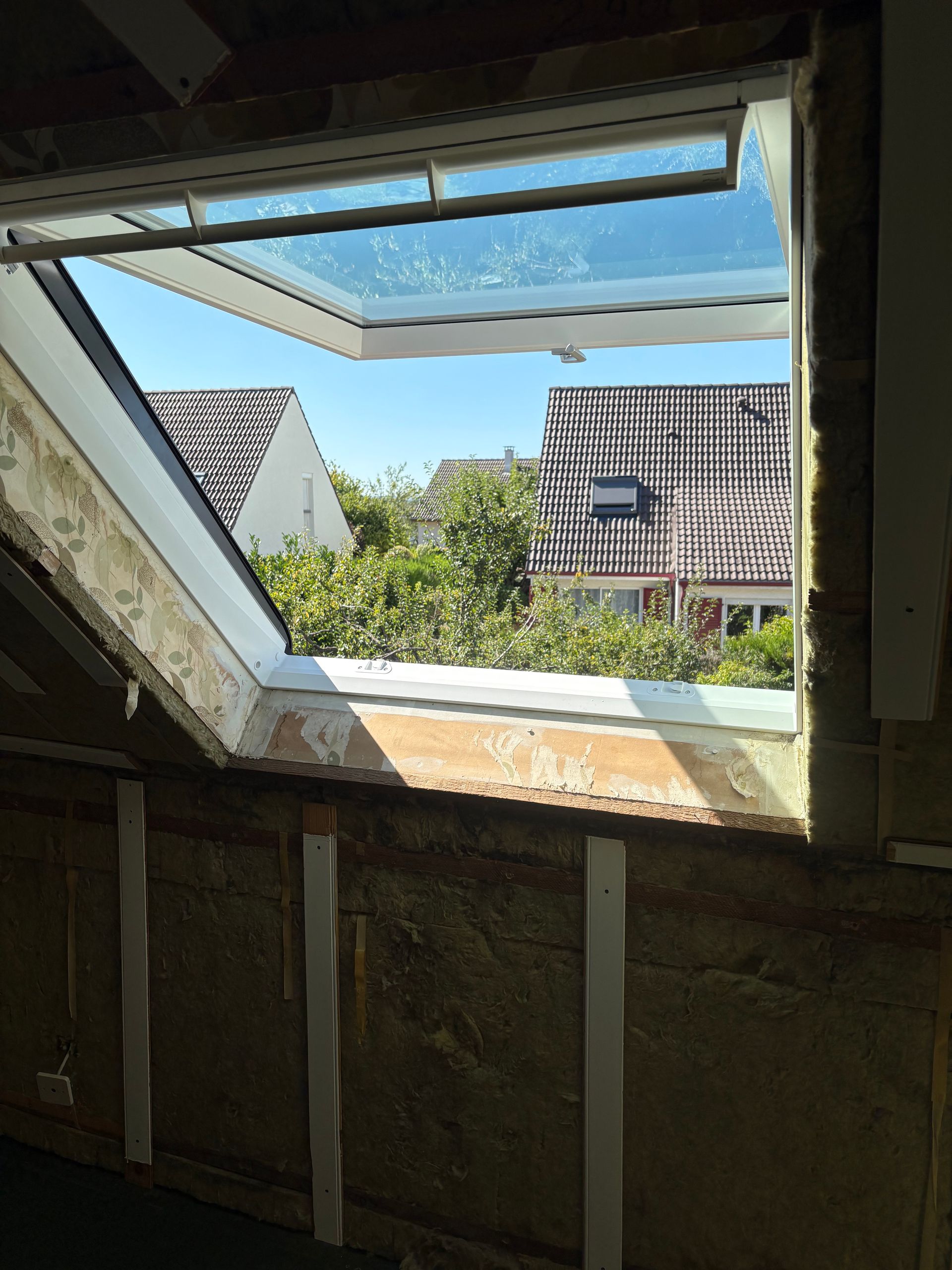Une lucarne ouverte dans une pièce dévoile une vue sur les toits, les arbres et le ciel bleu.