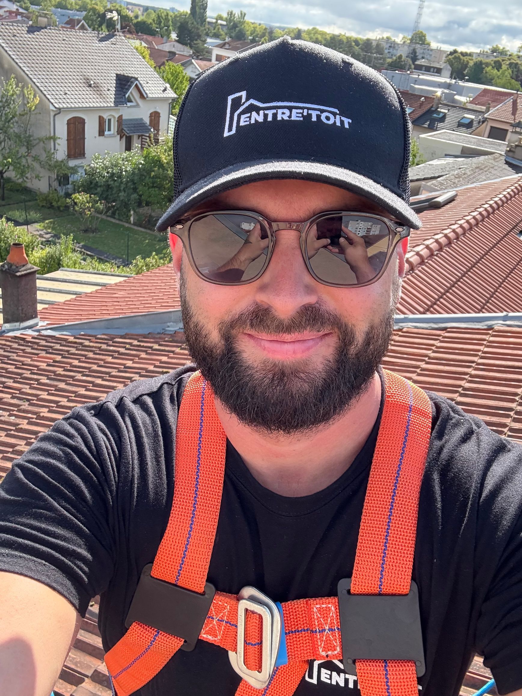 Un homme sur un toit, portant un harnais de sécurité, une casquette et des lunettes de soleil, sourit.