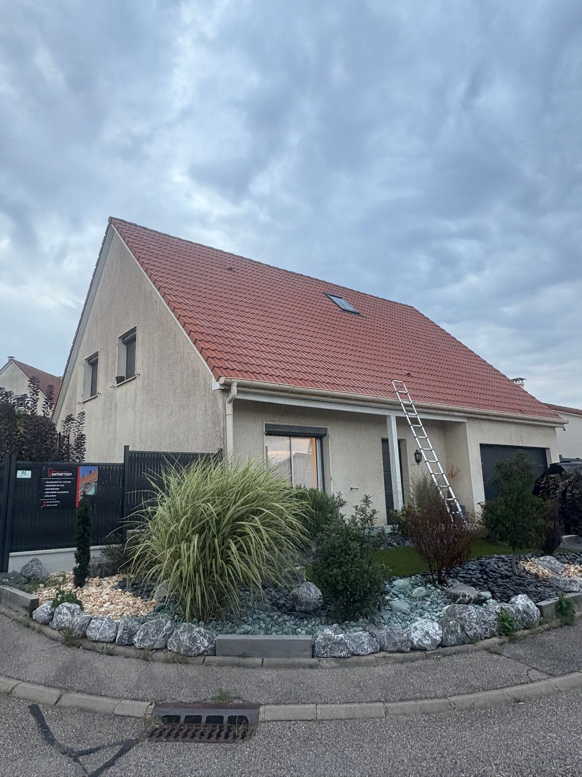 Maison au toit de tuiles rouges, aux murs beiges, avec une échelle appuyée contre le toit. Ciel couvert.