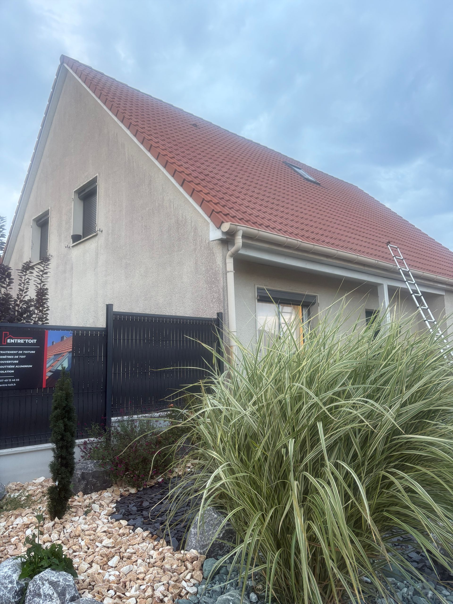 Maison beige au toit rouge, clôture sombre et gazon ornemental devant. Ciel nuageux.