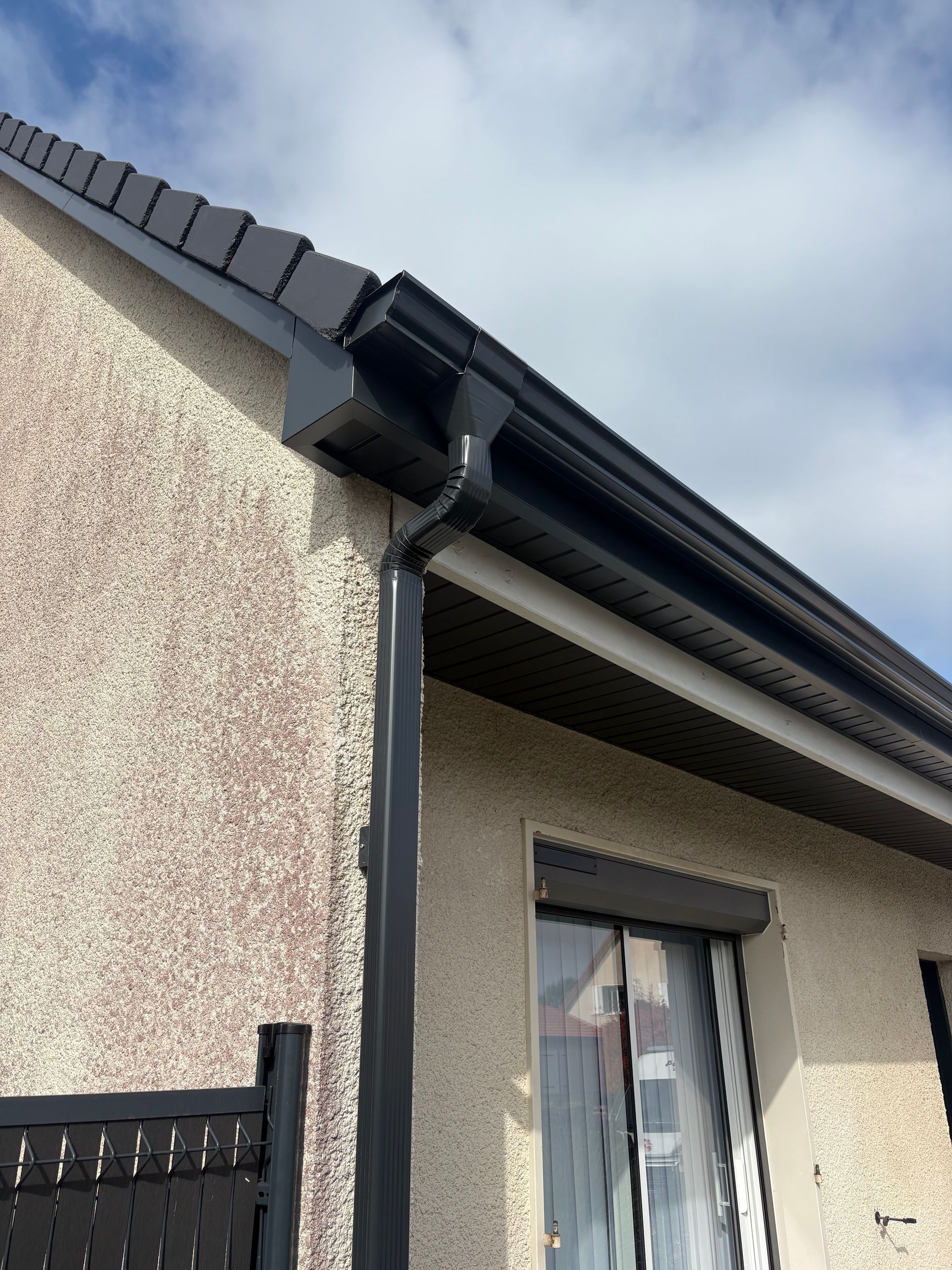Gouttières et descente pluviale noires sur une maison avec stuc texturé et une fenêtre avec store fermé.