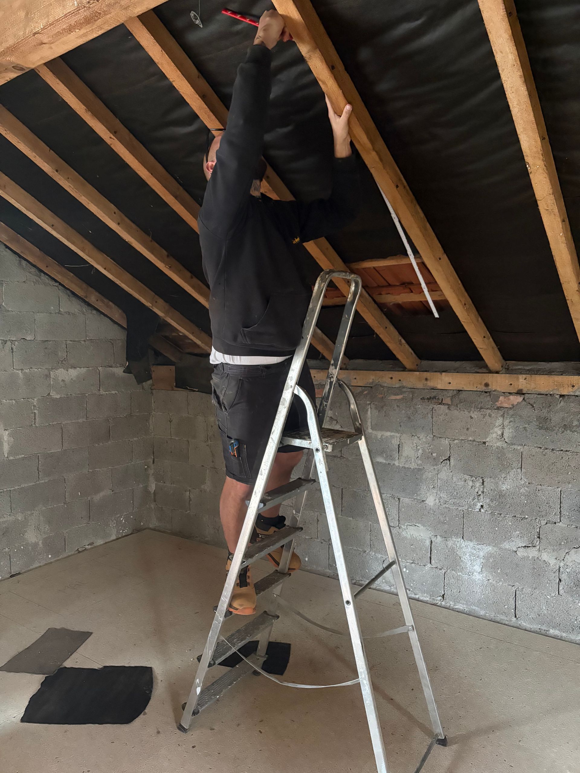Un homme sur une échelle travaille sur les chevrons du plafond d'une pièce inachevée.