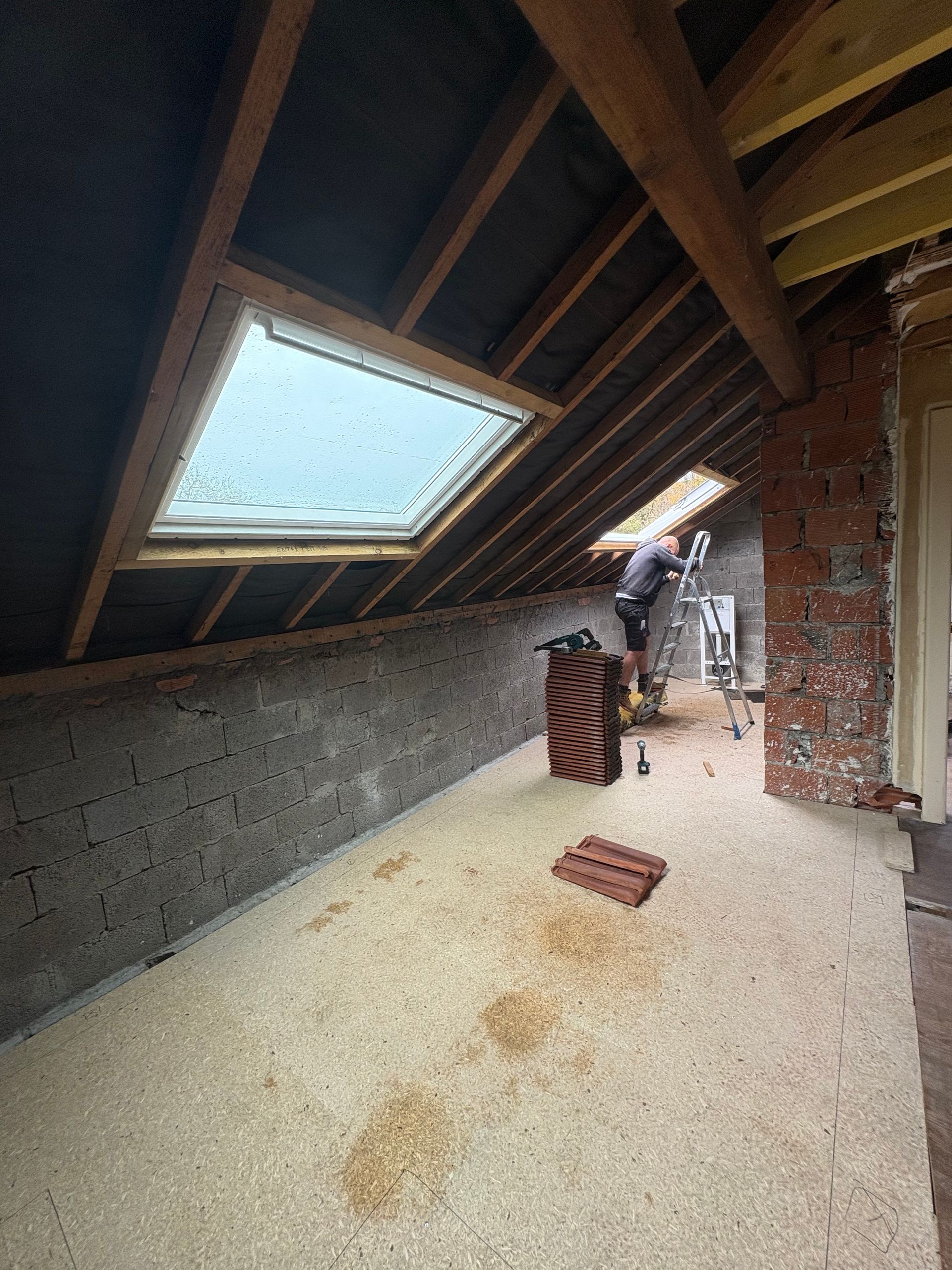 Intérieur d'un grenier avec un puits de lumière et une personne travaillant sur une échelle près d'un mur de briques.