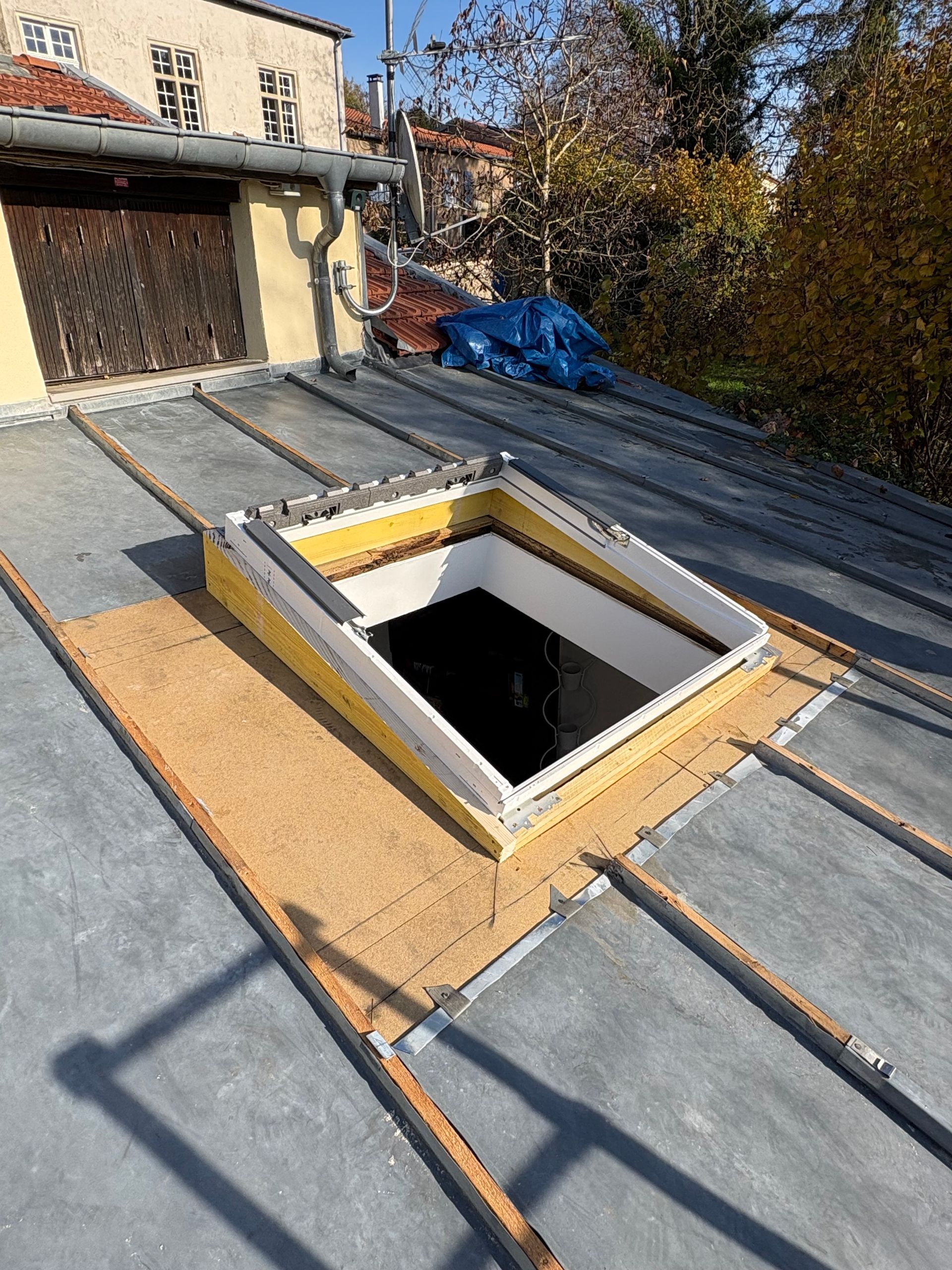 Puits de lumière sur un toit plat en métal, entouré de bois. Isolation jaune, bâtiment en arrière-plan.