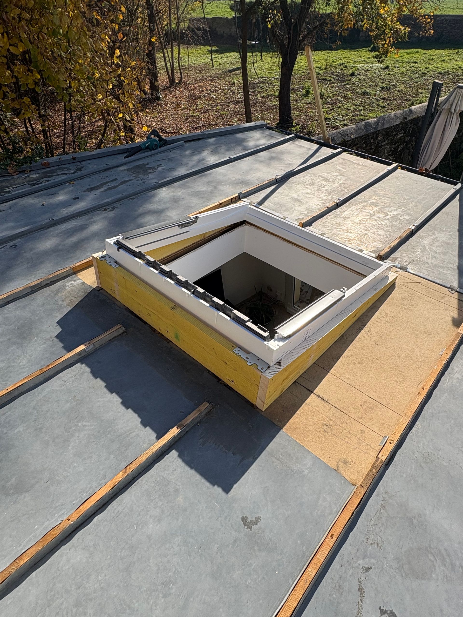 Installation d'un puits de lumière sur un toit plat, entouré d'un matériau de couverture gris et de supports en bois.