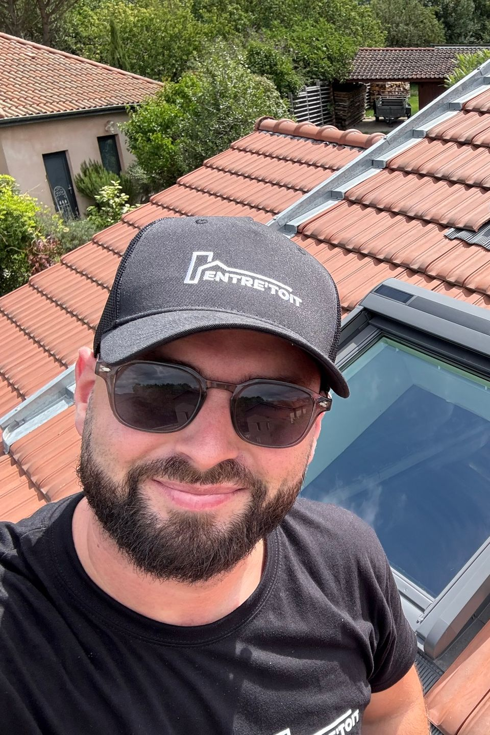 Un homme portant des lunettes de soleil et une casquette, souriant sur un toit de tuiles rouges, à côté d'une lucarne.