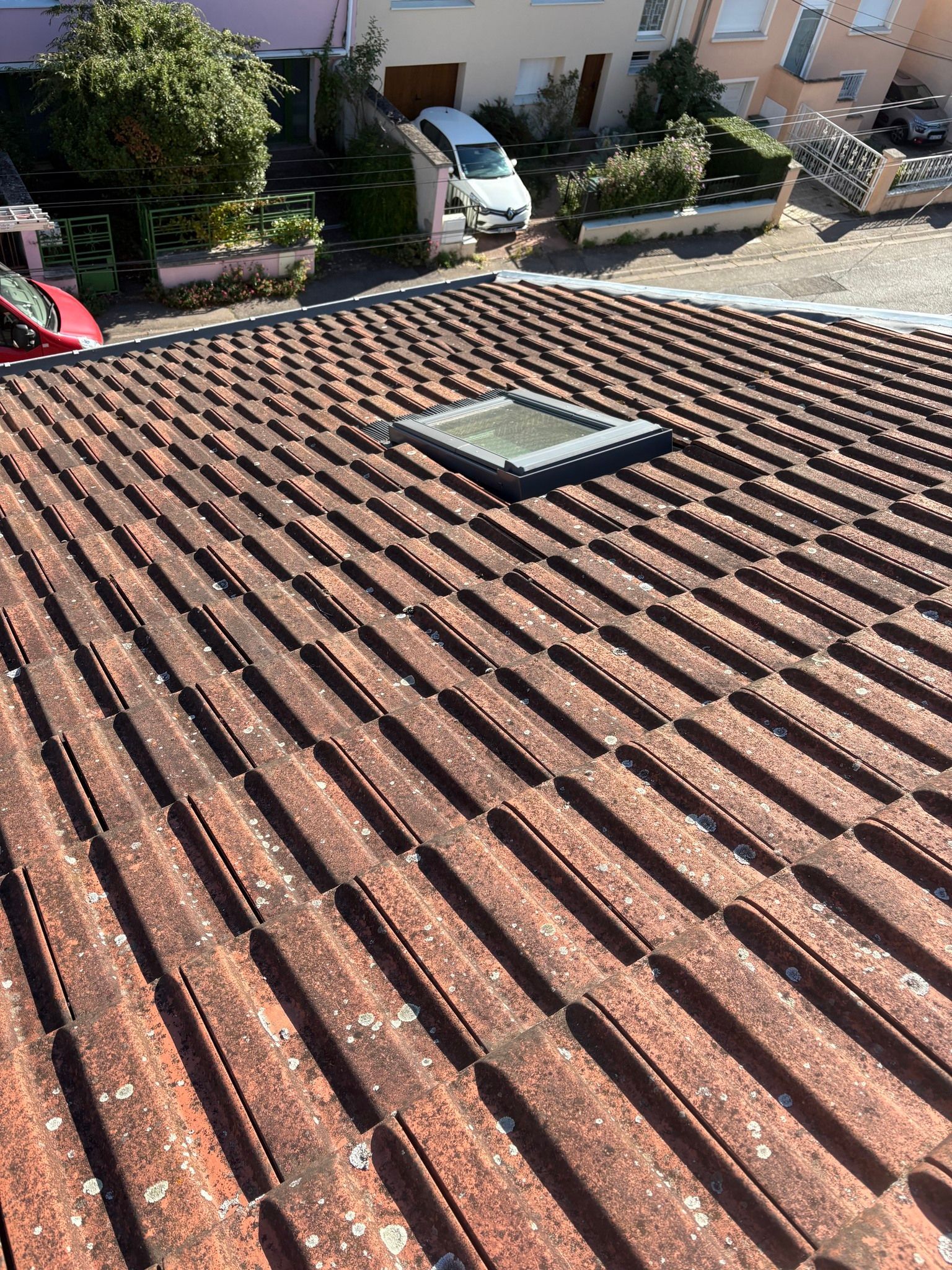Toit de tuiles rouges avec un puits de lumière, maisons et voitures en arrière-plan.