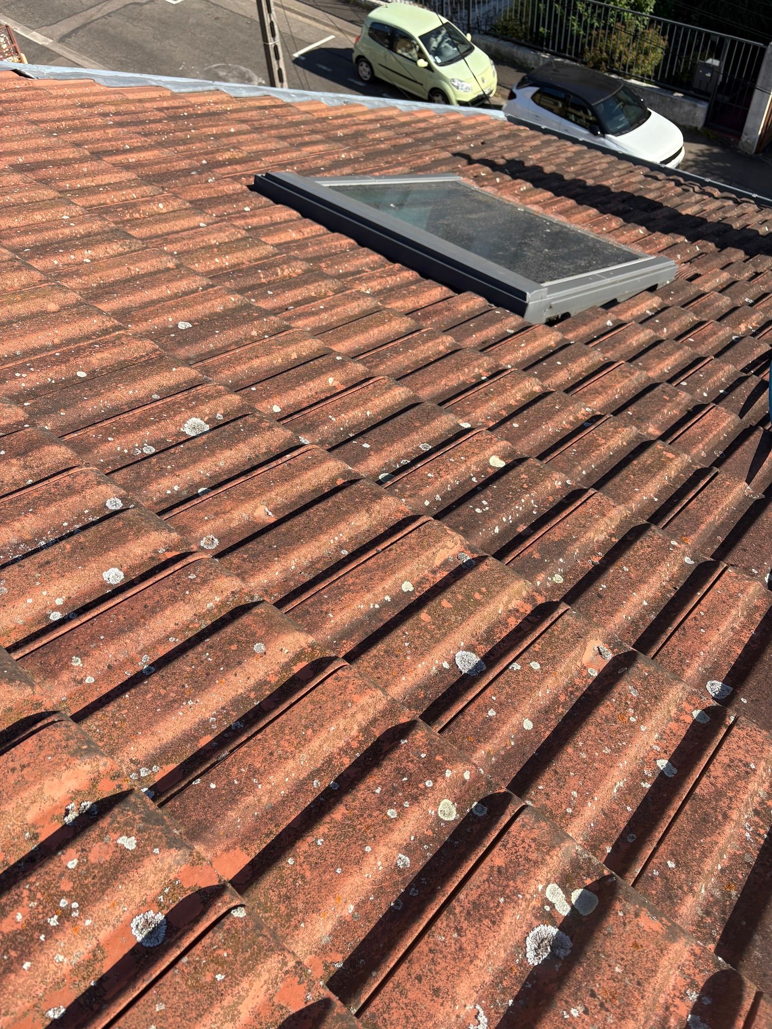 Gros plan sur un toit de tuiles rouges patiné par le temps, percé d'une lucarne ; voitures et route visibles en arrière-plan.