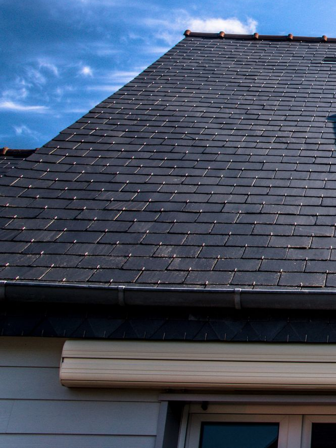 Toit d'ardoise gris foncé sur un ciel bleu, au-dessus d'une maison blanche aux volets beiges.