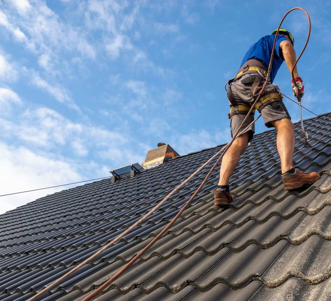 Couvreur sur un toit de tuiles sombres, portant un harnais de sécurité, sur fond de ciel bleu, au travail.