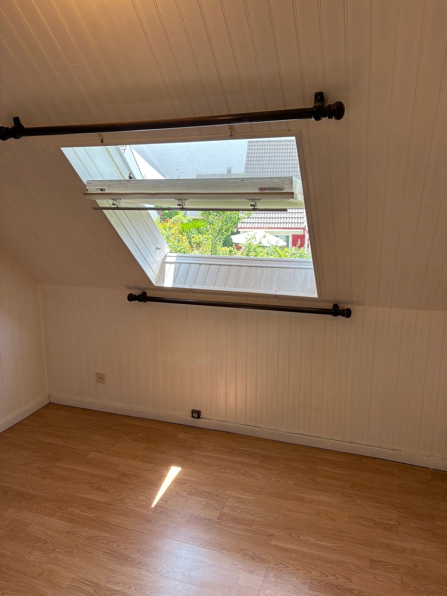 Photo intérieure d'une pièce mansardée avec un puits de lumière, des murs lambrissés blancs et un sol imitation bois.