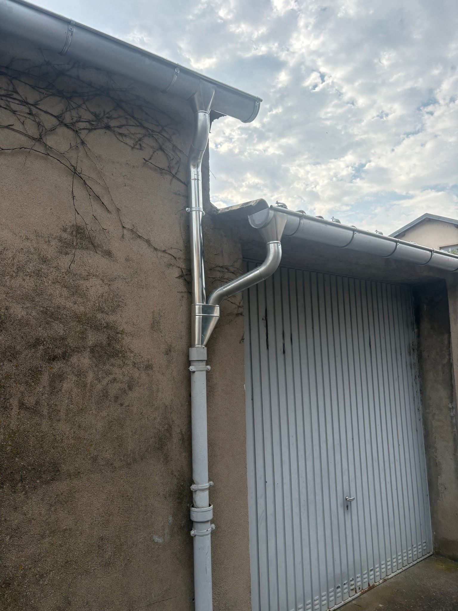 Gouttières et descente pluviale en métal gris sur un mur texturé beige, à côté d'une porte de garage assortie.