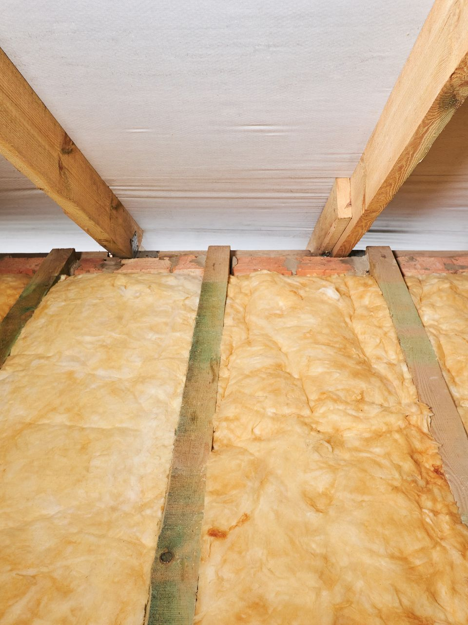 Isolation entre les poutres en bois dans un grenier non aménagé ; fibre de verre beige, bois brun et plafond blanc.
