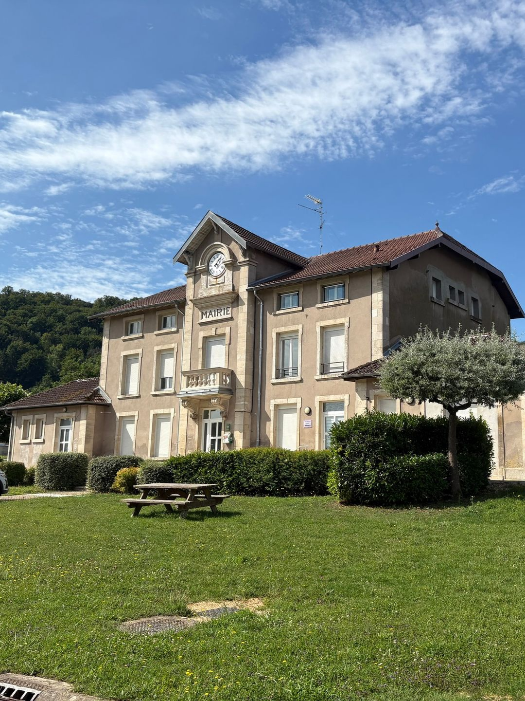 Grand bâtiment beige avec tour d'horloge, toit de tuiles rouges, pelouse verte, table de pique-nique et ciel bleu.