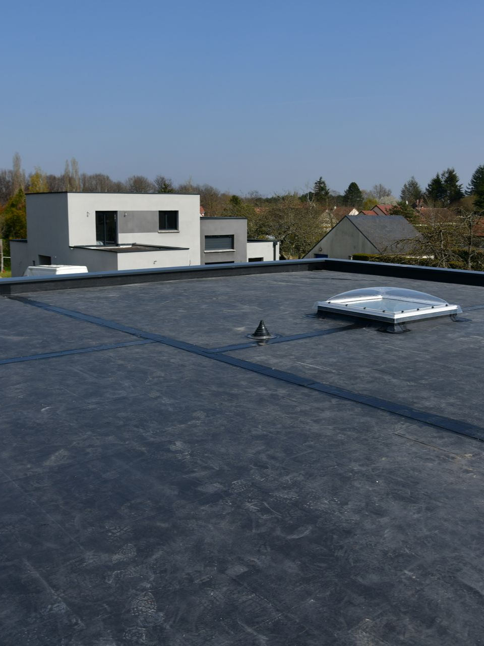 Toit plat noir avec un puits de lumière, un petit bâtiment en arrière-plan et un ciel ensoleillé.